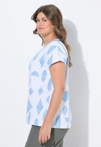 Camiseta blanca con patrones geométricos azules que incluyen líneas y formas. Mangas cortas y escote redondo. Modelo vistiendo pantalones verde oliva.