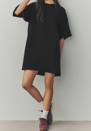 Personne portant une robe t-shirt noire oversize, des chaussettes blanches et des baskets bordeaux, debout avec une jambe croisée sur l'autre contre un mur uni.