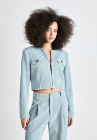 Svetle modrá cropped bunda s texturovaným vzorem, obsahující dvě přední kapsy, stříbrné kování a dlouhé rukávy, nošená s odpovídajícími kalhotami.