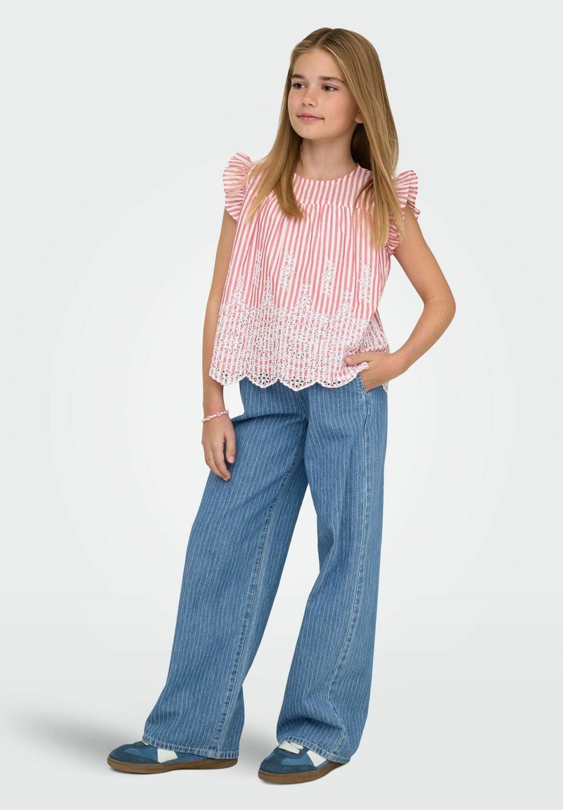 Jeune fille aux longs cheveux blonds, portant un haut rose à rayures avec des détails en dentelle, un jean large bleu à fines rayures et des baskets bleues, debout avec une main dans la poche.