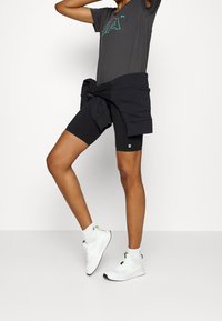 Shorts ajustados negros combinados con una camiseta gris que lleva un logo turquesa, y zapatillas deportivas blancas con acentos en neón. Calcetines blancos visibles.