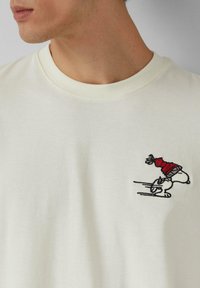 Cremfarbendes T-Shirt mit geripptem Rundhalsausschnitt; mit einer bestickten Figur mit rotem Hut auf der linken Brustseite. Weicher Stoff.