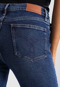 Mörkblå denimjeans med en åtsittande design. Har distinkta bakfickor med sömnadsdetaljer och en brun läderlogga.