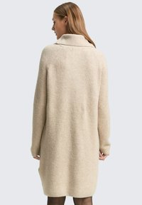 Beżowa sukienka swetrowa z wysokim kołnierzem, wykonana z miękkiego, teksturowanego materiału. Posiada długie rękawy i swobodny krój, sięgający powyżej kolan.