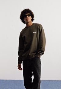 Sudadera verde oliva con cuello redondo y un pequeño logo blanco en el pecho, combinada con jeans oscuros de una tela texturizada.