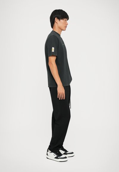 Sort bomulds T-shirt med korte ærmer, prydet med et grønt logo og et lille mærke på venstre arm, sammensat med sorte bukser og sneakers.