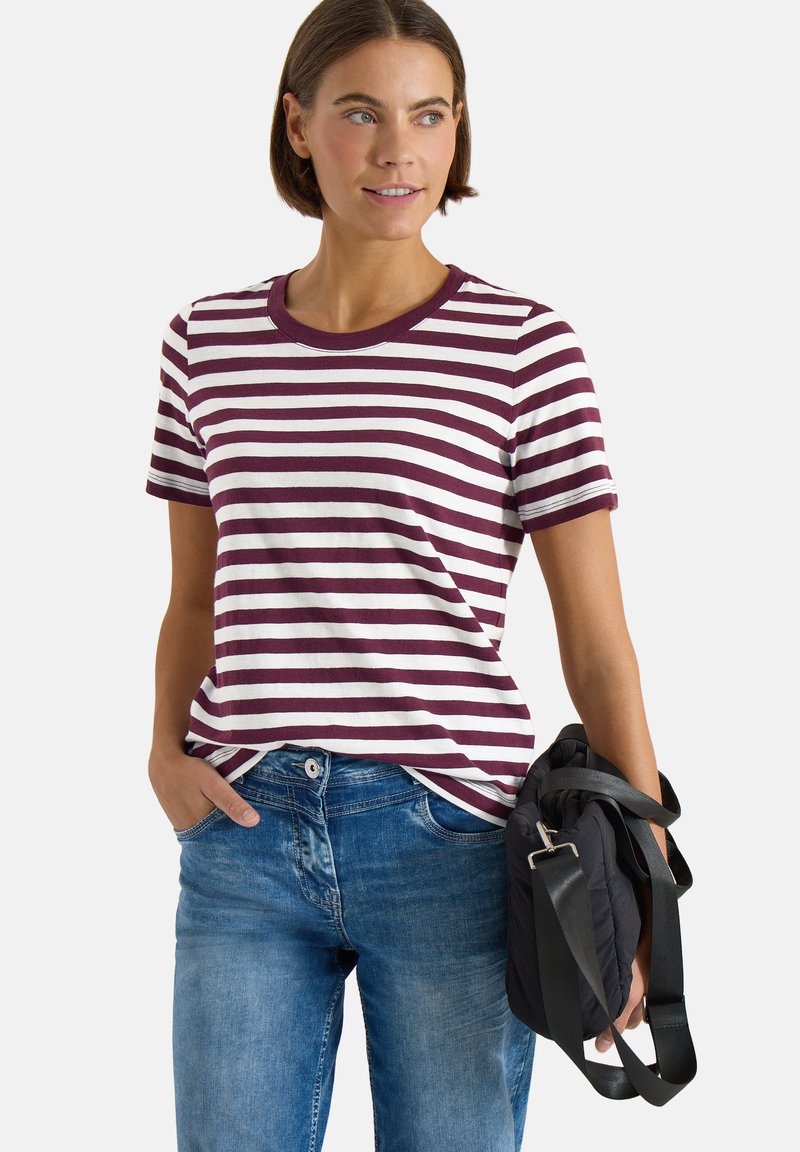 Frau trägt ein kurzärmeliges, bordeaux-weiß gestreiftes Shirt und blaue Jeans und hält eine schwarze Schultertasche mit verstellbaren Trägern.