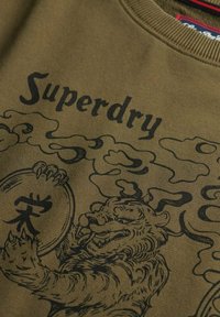 Tessuto verde oliva con una grafica nera di una creatura mitologica stilizzata che tiene un oggetto circolare e la parola "Superdry" sopra di essa.