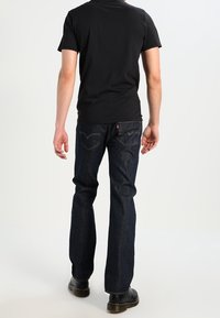 Tmavě modré džíny se střihem rovného nohavice, v kombinaci s upnutým černým tričkem, zobrazené zezadu. Černá obuv dokončuje outfit.