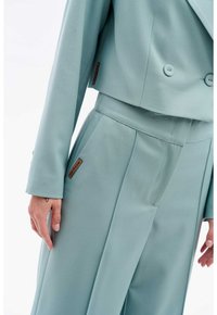 Costume bleu clair comprenant une veste cropped croisée et un pantalon à jambes larges. Tissu lisse avec un léger éclat, intégrant un accent de label de marque.