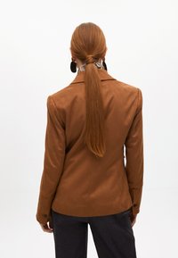 Hnědá nubuková sako s přesně padnoucím designem, dvěma knoflíky na manžetách a hladkou texturou. Výrazné jsou jeho strukturovaná ramena a úzký střih.