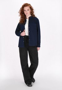 Chemise-jupe bleu marine avec un col et deux poches avant, associée à un pantalon noir ample et des chaussures noires. Texture lisse.