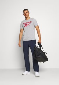 Camiseta cinza da Reebok com logotipo vermelho, calças de treino azul marinho e sapatos brancos; segurando uma mala de duffle preta com alças ajustáveis.