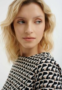 Blusa con un motivo geometrico in nero, bianco e beige, con collo a barca e texture di tessuto morbido. Capelli biondi leggermente disordinati in vista.