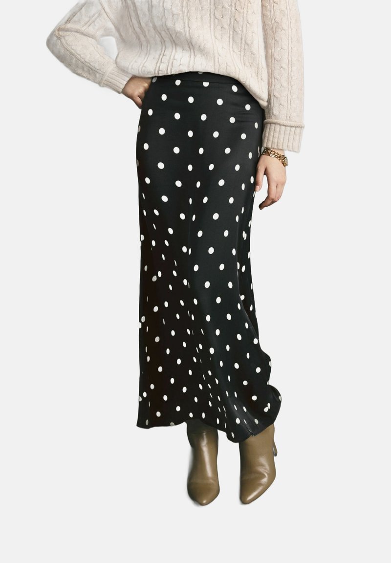 Personne portant une longue jupe noire à pois blancs, un pull en maille crème, un bracelet doré et des bottines taupe sur fond blanc.