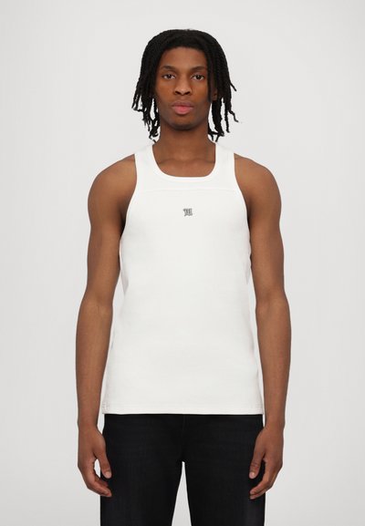 Hvid bomulds tanktop med ribbet tekstur, brede stropper og en lille logo detalje på brystet. Båret med mørke jeans.
