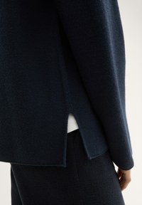 Pull en tricot bleu marine foncé avec une fente sur le côté, présentant un tissu texturé et un col rond, associé à des pantalons assortis.