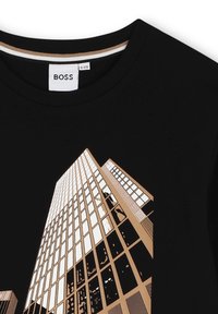 T-shirt noire avec un imprimé graphique représentant des bâtiments urbains en beige et blanc, avec des lignes architecturales et des reflets. Étiquette BOSS visible.