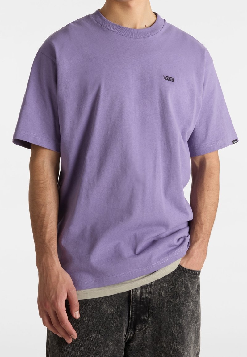 Homme portant un t-shirt Vans violet clair à manches courtes et un jean noir délavé, debout avec une main dans la poche.
