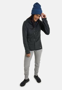 Black fleece jacket with zip closure, grey pants, and blue knit beanie. Features include a front pocket and a fitted design. Black footwear shown.