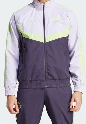 Hombre vistiendo un chándal Adidas azul violeta y amarillo claro con chaqueta con cremallera y pantalones a juego, con parches de logo y tres franjas en las mangas.