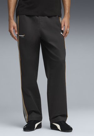 Man in zwarte McLaren Racing-broek met gouden zijstrepen en zwarte Puma-sneakers tegen een effen grijze achtergrond.