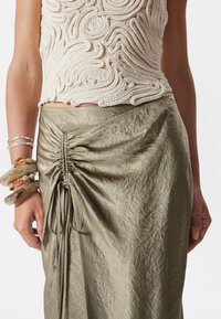 Beige strukturierte Bluse mit erhobenen, seilartige Mustern, kombiniert mit einem glänzenden olivgrünen Rock, der seitliche Raffung und eine Bindeschleife aufweist.