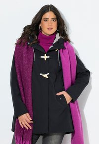 Kvinna med mörkt vågigt hår bär en svart kappa med knappkrokar, en ljuslila polotröja och en tvåfärgad lila scarf över gråa byxor.