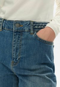 Blå denimjeans med en flätad midja, sidoficka och kontrasterande orange sömnad. Tyget är strukturerat och något slitet.