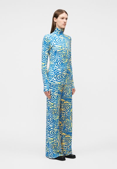 Γυναίκα που φοράει ένα τατουλίν με μοτίβο σε μπλε και κίτρινο χρώμα και φαρδιά παντελόνια, στέκεται μπροστά σε ένα απλό λευκό φόντο.