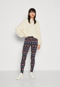 Tights med en mørkeblå bakgrunn med et festlig mønster, inkludert pepperkakefigurer, snøfnugg og tekst. Brukt med en stor kremfarget genser.
