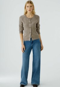 Khakifarbener Strickcardigan mit Rundhalsausschnitt, Knopfleiste und zwei Taschen. Kombiniert mit weit geschnittenen blauen Jeans und dunklen Schuhen.