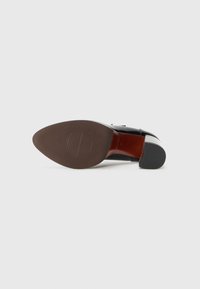 Chaussure en cuir noir avec un bout pointu, une finition lisse et une semelle en caoutchouc marron présentant un motif de grip circulaire.