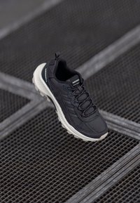 Schwarzer Mesh-Sportschuh mit strukturiertem Obermaterial, ausgestattet mit Schnürung, gepolsterter Zwischensohle und kontrastierender cremefarbener Außensohle.