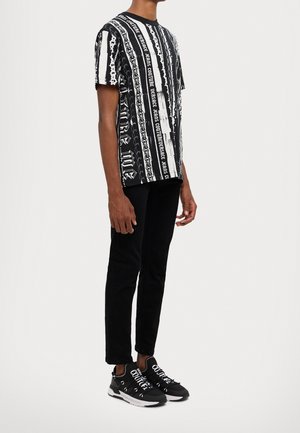 Schwarzes und weißes Grafik-T-Shirt mit kurzen Ärmeln, Text- und Kettenmustern, kombiniert mit schwarzer Hose und schwarzen Sneakers mit weißen Akzenten.