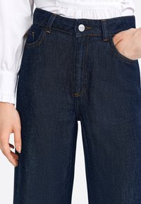 Dark blue denim jeans with a straight leg design, featuring orange stitching, front pockets, and a silver button closure at the waist.