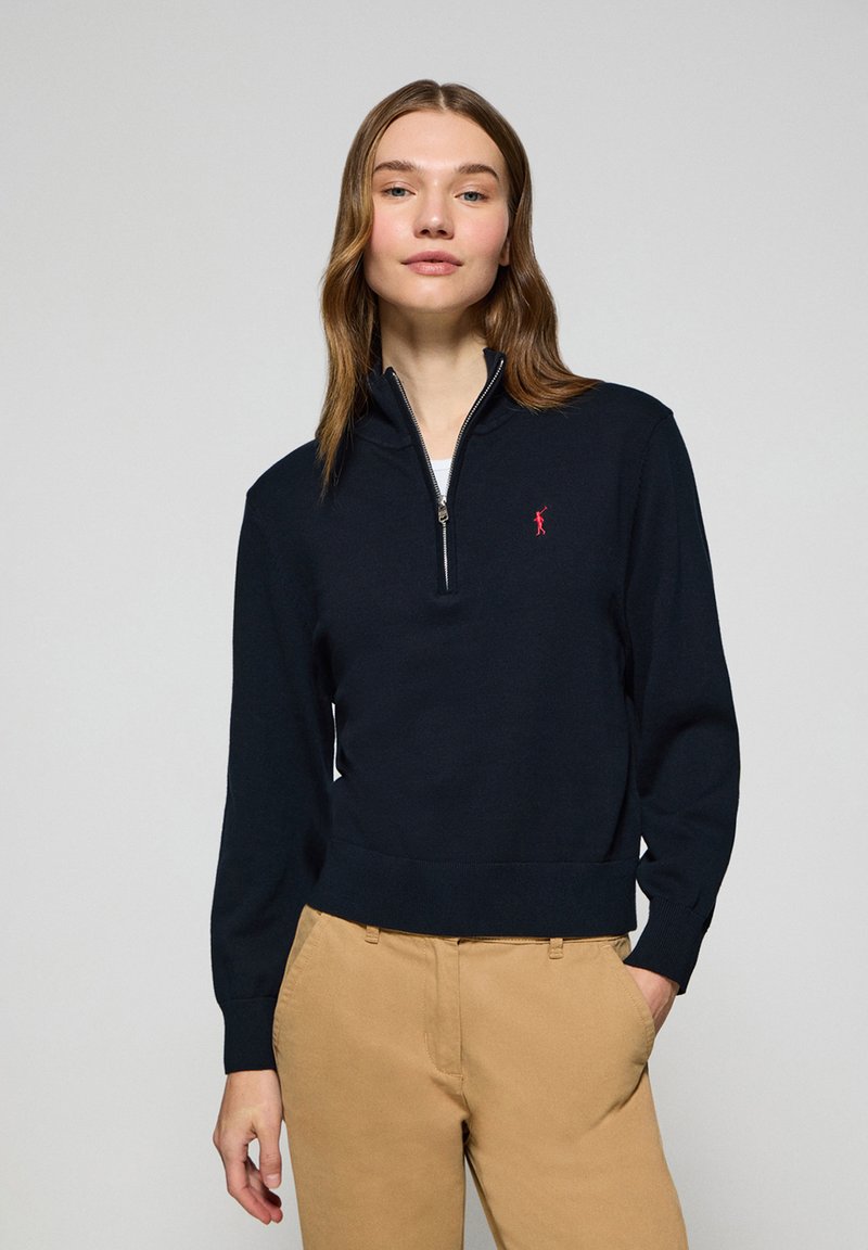 Pull-over zippé marine avec manches longues, poignets et ourlet côtelés. Arbre d'un logo rouge sur la poitrine gauche. Porté sur une chemise blanche et un pantalon beige.
