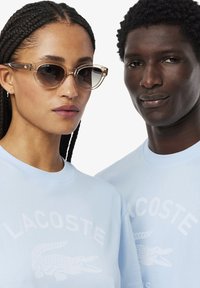 Gafas de sol transparentes con un marco marrón claro, usadas por una persona con una camisa azul clara que presenta un logo de cocodrilo blanco, al lado de otro modelo.