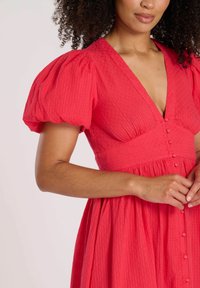 Röd klänning med puffärmar i texturerad tyg, med djup V-ringning och knapppanel vid midjan, samt en flödande kjol.