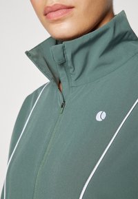Grønn sportsjakke med høy krage, glidelås foran og kontrastfarget hvit piping. Har et lite logo på brystet og en glatt tekstur.