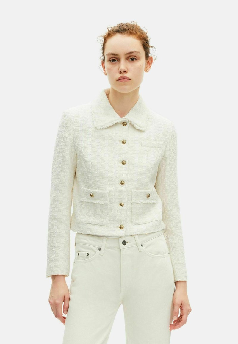 Femme portant une veste blanche texturée avec des boutons dorés et deux poches avant, associée à un pantalon blanc taille haute.