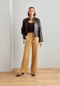Bruine leren jas met hoge kraag, zwarte kanten top, beige wijde broek en donkere hakken. Glad textuur; ontspannen pasvorm.