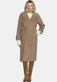 Beżowy, wełniany płaszcz z paskiem, dużymi klapami i guzikiem na mankietach. Połączony z beżowymi, wysokimi na kolana botkami. Widoczne teksturowane rękawy.