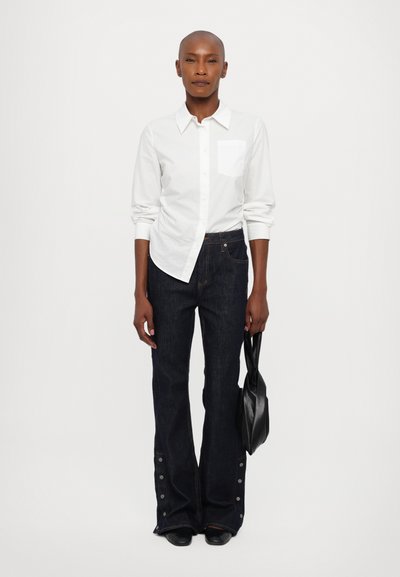 Hvit langermet skjorte med lomme, kombinert med mørk denim flared jeans med trykknappdetaljer ved ermene. Modellen holder en svart veske.