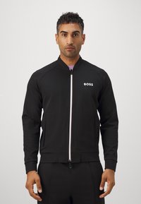Svart zip-up jacka med hög krage, tillverkad av slät tyg. Har en vit logotyp på övre vänster sida och framfickor. Skräddarsydd passform.