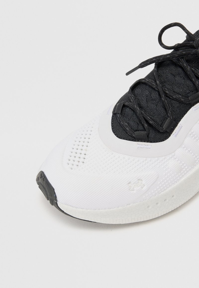 Zapatilla deportiva en blanco y negro con parte superior de malla, detalles perforados, cordones texturizados en negro y suela de goma. Presenta un logo de Under Armour.