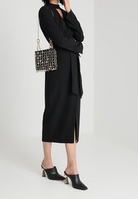 Mujer con un vestido midi negro con una abertura frontal, tacones de tiras negros y un bolso de cadena de patrón en blanco y negro.