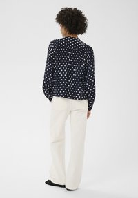 Blouse marine à pois avec manches longues et détail froncé à l'arrière, associée à un pantalon ample couleur crème et des chaussures noires.