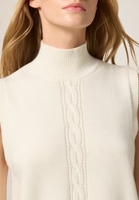 Suéter sin mangas de color crema con un cuello alto acanalado y un patrón de trenzado central, que presenta una textura suave y una estructura de punto uniforme.
