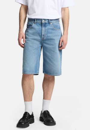 Shorts en denim bleu clair, arrivant au genou, avec une coupe décontractée. Comprend des passants de ceinture, cinq poches et un ourlet double couture. Associés à des chaussures noires.